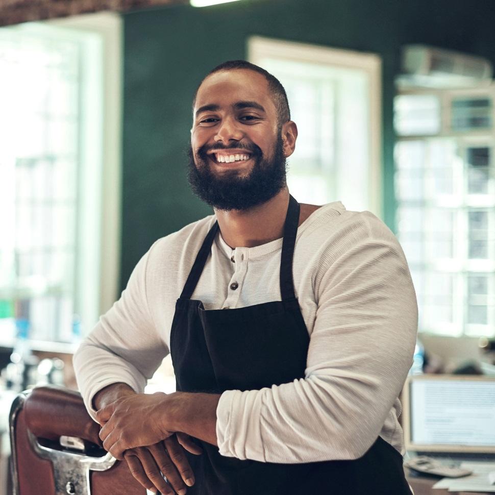 Smiling person in black apron and white shirt in a bright cafe or restaurant setting.